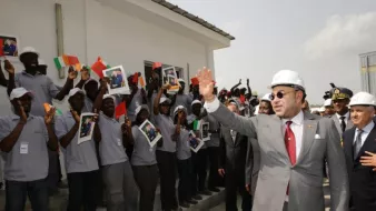 L'image montre un groupe de personnes, probablement des travailleurs ou des étudiants, levant des affiches avec des photos tout en saluant une personne qui semble être un dirigeant. Ce dernier porte un casque de sécurité et coûte en costume, indiquant une visite d'inspection ou une inauguration. Les personnes en arrière-plan sont toutes habillées de manière uniforme et semblent enthousiastes, créant une atmosphère de célébration. L'environnement est extérieur, avec un bâtiment en arrière-plan.
