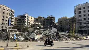 L'image montre une scène de destruction urbaine, où plusieurs bâtiments sont partiellement effondrés et en ruines. On peut voir des décombres éparpillés au sol, tandis qu'une personne à moto passe devant. Le ciel est dégagé, laissant transparaître un jour ensoleillé, contrastant avec le désastre visible. La scène évoque un fort sentiment de désolation et de dégâts, typique de zones touchées par des conflits ou des catastrophes.
