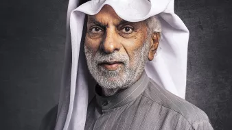 L'image montre un homme âgé avec une barbe grise, portant un vêtement traditionnel de style arabe, typiquement un thobe gris avec des rayures. Il a également un agal noir et un ghutrah blanche, qui est un couvre-chef traditionnel. L'arrière-plan est sombre, ce qui met en valeur son expression sage et son apparence traditionnelle.