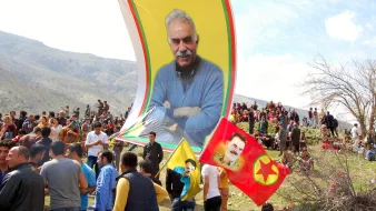 L'image montre une grande réunion en plein air, probablement lors d'une célébration ou d'un événement politique. On peut voir un grand drapeau avec le portrait d'un homme, entouré de plusieurs drapeaux aux couleurs vives, notamment du jaune et du rouge. De nombreuses personnes sont rassemblées et semblent animées, certaines brandissant des drapeaux. En arrière-plan, des montagnes et des arbres se dressent, suggérant un cadre naturel. L'atmosphère semble festive et engagée.