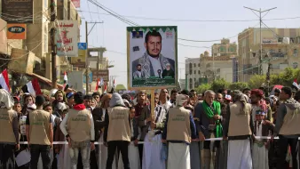 تظهر في الصورة مجموعة من الأشخاص مجتمعين في مظاهرة أو تجمّع شعبي. في الخلفية، يُرفع لافتة كبيرة تحمل صورة لأحد الشخصيات، ويبدو أن المتظاهرين يرتدون ملابس متشابهة. تعكس الأعلام الموجودة في الصورة انتماءات وطنية، كما يظهر أن هناك تفاعلًا وحماسًا بين المشاركين. الجو يعكس نوعًا من الاحتشاد والتضامن حول قضية معينة.
