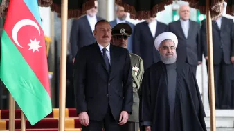 En la imagen se pueden ver a dos líderes de países que están de pie juntos. A la izquierda, hay una bandera de Azerbaiyán. Uno de los líderes lleva un traje negro y corbata, mientras que el otro viste una indumentaria tradicional oscura. Detrás de ellos, hay un grupo de personas que parecen ser miembros de la seguridad o funcionarios, y en el fondo se observan algunos edificios. La escena parece ser un acto formal o una reunión oficial.