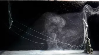The image showcases a mesmerizing underwater scene featuring swirling currents and bubbles. The foreground has a smooth, almost glass-like surface with a hint of foam, while the background features abstract formations that resemble smoke or flowing water. Thin, delicate tendrils or strings extend across the scene, creating an ethereal atmosphere. The overall color palette is dominated by dark tones, emphasizing the luminous qualities of the water and creating a sense of depth and motion.