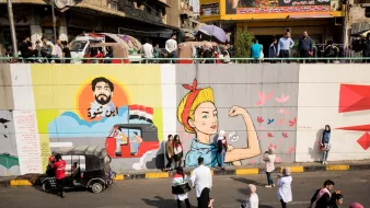 L'immagine mostra un murale su un grande muro divisorio, con diverse sezioni artistiche. A sinistra, c'è un ritratto di un uomo con una barba, che sembra essere ispirato a un personaggio storico o culturale, accompagnato da elementi grafici che richiamano il paesaggio urbano. A destra, c'è l'immagine di una donna in posa iconica, che rappresenta la forza e l'emancipazione, con dettagli colorati e dinamici. In basso, si osserva un gruppo di persone che passeggiano lungo la strada, creando un contrasto tra l'arte e la vita quotidiana. La scena riflette una vivace interazione tra arte, cultura e comunità.