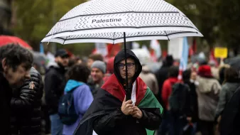 En la imagen, se puede ver a una persona que sostiene un paraguas con la palabra "Palestine" escrita en él. La persona lleva una bandera de Palestina sobre los hombros y parece estar en una manifestación o protesta. Alrededor se pueden observar otras personas, algunas con paraguas y banderas, en un ambiente cargado de emociones y solidaridad. El clima parece nublado o lluvioso, lo que añade a la atmósfera del evento.