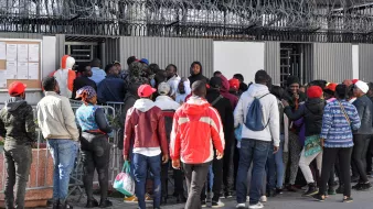 L'image montre un groupe de personnes rassemblées devant une clôture avec des barbelés. Elles semblent attendre ou essayer d'entrer dans un bâtiment. Certaines personnes portent des vêtements colorés, comme des vestes et des chapeaux rouges. Il y a une certaine densité dans la foule, suggérant une situation de rassemblement ou de file d'attente. Le mur en arrière-plan comporte des affichages et il y a une ambiance de tension ou d'importance autour de l'endroit où se trouvent les personnes.