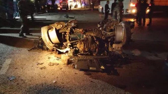 The image depicts a severely damaged vehicle that has been overturned. The car's chassis and engine are exposed, showing significant destruction, with debris scattered around the area. The scene appears to be dark, possibly at night, with some emergency responders or bystanders in the background, indicating that it may have been the site of an accident. The overall context suggests a serious incident involving the vehicle.