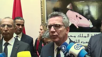 L'image montre un groupe de personnes lors d'une conférence de presse ou d'un événement officiel. Au premier plan, un homme s'exprime devant des caméras et des micros, entouré de plusieurs autres individus qui semblent attentifs. En arrière-plan, on peut voir un portrait ou une image représentant des mains avec des symboles significatifs, ainsi qu'un drapeau. L'atmosphère semble formelle, probablement liée à des discussions d'ordre politique ou diplomatique.