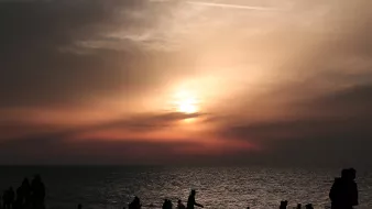 L'immagine mostra una scena al tramonto sulla spiaggia. Il cielo è dipinto di tonalità calde, con sfumature di arancione e rosa, mentre il sole si avvicina all'orizzonte. In primo piano, si vedono diverse silhouette di persone che camminano lungo la riva, alcune sedute o accoccolate sulla sabbia. La superficie dell'acqua riflette i colori del cielo, creando un'atmosfera calma e suggestiva.