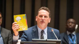 L'image montre un homme s'exprimant lors d'une réunion. Il tient un document ou un carnet dans sa main droite et semble faire un discours. L'homme est vêtu d'un costume sombre avec une cravate. Derrière lui, on aperçoit d'autres personnes assises, dont l'une semble attentivement écouter. Un panneau avec le nom "PIERRE KRAHENBUHL" est visible sur la table. L'ambiance semble formelle et professionnelle.