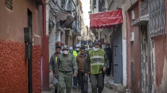 الصورة تظهر زقاقًا ضيقًا في إحدى المدن، حيث يسير مجموعة من الأشخاص. يرتدي بعضهم زيًا رسميًا، مما يشير إلى أنهم من عناصر الأمن أو الشرطة، ويبدو أنهم يقومون بدورية أو لجنة تفتيش. تظهر خلفهم مبانٍ قديمة وممرات ضيقة، مع بعض العلامات التجارية والستائر الملونة. الأجواء توحي بنشاط وحركة في المكان.