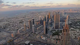 The image depicts a stunning aerial view of a sprawling cityscape, likely a metropolitan area with modern architecture. Skyscrapers dominate the skyline, showcasing a mix of designs and heights. Below, intricate road systems weave through the landscape, indicating a bustling urban environment. In the background, the coastline meets the ocean, with a hint of the horizon showcasing a picturesque sunset. The combination of urban infrastructure and natural beauty creates a striking contrast, highlighting the vibrancy of the city.
