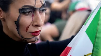 L'image montre une femme avec un maquillage artistique qui représente une carte. Elle a des lignes noires lisses sur son visage qui dessinent une forme géographique, accompagnées d'une larme rouge. Sa bouche est accentuée par un rouge à lèvres également rouge. Elle tient un drapeau, probablement national, avec des couleurs distinctives. L'arrière-plan semble flou, avec d'autres personnes en mouvement, suggérant un rassemblement ou une manifestation.