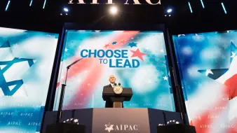 L'image montre un homme sur scène, probablement un orateur, devant un public. Il se tient derrière un pupitre avec le logo d'AIPAC, une organisation pro-israélienne. En arrière-plan, il y a un grand écran affichant le slogan "CHOOSE TO LEAD" (Choisissez de diriger), entouré de motifs étoilés. L'ambiance semble formelle et dédiée à un discours important, probablement lors d'une conférence ou d'un événement lié à AIPAC.