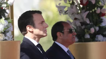 این تصویر دو مرد را در کنار هم نشان می‌دهد. شخصی در سمت چپ با موهای تیره و لباس رسمی مشکی، در حالت تفکر و جدی ایستاده است. در سمت راست، مرد دیگری با عینک آفتابی و لباس رسمی به رنگ تیره، در کنار او قرار دارد. پس‌زمینه شامل گل‌های سفید و عناصر طبیعی است که به تصویر زیبایی و شکیل بودن می‌بخشد.