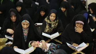 در تصویر گروهی از زنان با چادرهای سیاه در حال مطالعه قرآن دیده می‌شوند. بعضی از آن‌ها در حال خواندن هستند و توجه عمیقی به کتاب دارند. همچنین، یک کودک در آغوش یکی از زنان خوابیده است. به‌طور کلی، فضایی معنوی و پر از تمرکز در این تصویر وجود دارد.