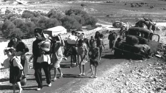 L'image montre un groupe de personnes marchant le long d'une route, probablement en situation de déplacement ou d'exode. On peut voir des adultes portant des enfants et des charges, tandis que des enfants marchent également. À l'arrière-plan, des véhicules abandonnés et un paysage aride s'étendent, suggérant une situation difficile. L'atmosphère semble grave et chargée d'émotion, témoignant des épreuves que traversent ces personnes.