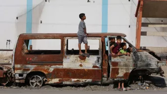 L'immagine mostra un gruppo di bambini che interagiscono attorno a un veicolo abbandonato e arrugginito. Uno dei bambini è in piedi sulla parte superiore di un furgone, mentre altri si trovano all'interno e intorno al veicolo. Lo sfondo evidenzia un edificio deteriorato, suggerendo un ambiente urbano trascurato. L'atmosfera sembra essere quella di un'area in difficoltà, ma i bambini sembrano divertirsi nonostante le condizioni.