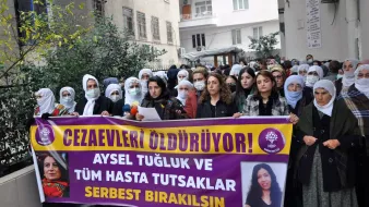 En la imagen, un grupo de personas se encuentra reunido en una manifestación. Hay mujeres con pañuelos en la cabeza, y algunas de ellas sostienen pancartas que expresan mensajes en apoyo a Aysel Tuğluk y a otros prisioneros enfermos. El ambiente parece ser de protesta, con un enfoque en la liberación de detenidos. Se observa una edificación al fondo, que sugiere que la manifestación se lleva a cabo frente a un edificio público o institucional.