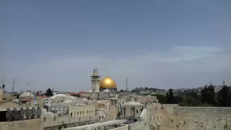L'immagine mostra una vista panoramica di una città con una predominante cupola dorata, che è probabilmente il Dôme della Roccia, un noto sito religioso a Gerusalemme. Intorno ci sono edifici con tetti piatti e una torre, mentre il cielo appare sereno e leggermente nuvoloso. Gli alberi e le colline sullo sfondo aggiungono profondità alla scena.