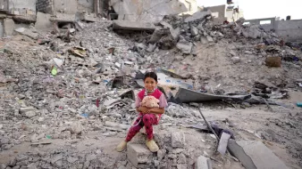 L'immagine ritrae una bambina seduta su un cumulo di macerie, circondata da un paesaggio distrutto. Il suo sguardo è diretto verso la fotocamera, mentre tiene un peluche tra le braccia. Indossa un pigiama a pois rosa e sembra essere in un ambiente devastato, con edifici in rovina sullo sfondo. La scena trasmette una forte emozione, evidenziando la vulnerabilità e la resilienza della giovane ragazza in un contesto difficile.