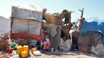 L'image montre une habitation informelle construite avec des matériaux comme des bâches en plastique et des morceaux de toile. À l'avant, on peut voir une femme assise dans l'ombre de la structure, entourée de divers objets et contenants, probablement utilisés pour la vie quotidienne. L'environnement semble un peu désordonné, avec des débris et des matériaux épars autour de l'habitation. Le ciel est dégagé, ce qui donne une impression de chaleur.