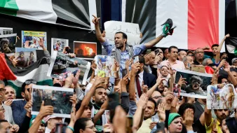 L'immagine mostra una folla di persone radunate in una manifestazione. Gli individui alzano cartelli e fotografie, esprimendo chiaramente le loro emozioni e il loro supporto per una causa. Alcuni di loro indossano sciarpe e altri simboli, mentre l'atmosfera sembra essere intensa e piena di passione. Sullo sfondo, ci sono bandiere che suggeriscono un contesto politico o sociale significativo.