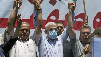 الصورة تُظهر مجموعة من الرجال في تجمع حاشد، حيث يرفع بعضهم أيديهم في إشارة للتضامن أو الاحتجاج. يرتدي أحد الأشخاص في المنتصف قناعاً للوجه، مما يشير إلى أنه قد يكون في سياق صحي معين. خلفهم يوجد لافتة كبيرة مكتوب عليها نص باللون الأحمر. الجو العام يوحي بالتعبير عن موقف أو رسالة معينة.