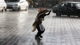 Nell'immagine si vede un bambino che corre sotto la pioggia intensa. Il ragazzo tiene un grande contenitore in una mano e si protegge dalla pioggia con un pezzo di stoffa. Sullo sfondo, ci sono diverse auto e una strada bagnata, mentre le gocce d'acqua creano un'atmosfera drammatica e vivace. La scena trasmette una sensazione di urgenza e resilienza, evidenziando il contrasto tra il bambino e le avversità del tempo.