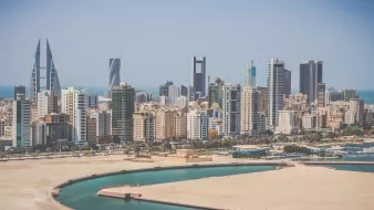 L'image montre un paysage urbain moderne avec une vue sur une ville côtière. On peut voir de nombreux immeubles de grande hauteur, représentant une architecture contemporaine, avec des designs variés allant des structures angulaires aux façades en verre. À l'arrière-plan, la mer est visible, ainsi qu'une baie ou un port. Le ciel est clair et ensoleillé, ce qui donne une impression de chaleur et de vivacité à la scène. La composition évoque une ambiance urbaine dynamique, typique de villes en développement.