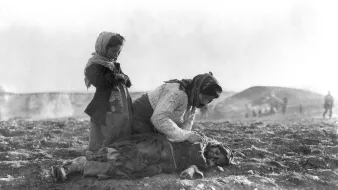 L'immagine mostra una scena drammatica e toccante. Una donna si trova inginocchiata su un terreno aperto, mentre si prende cura di un bambino disteso a terra, visibilmente in difficoltà. Un'altra bambina, in piedi accanto a loro, osserva con espressione seria. La scena è in un contesto desolato, con uno sfondo che suggerisce un ambiente di guerra o di sofferenza, evidenziando la vulnerabilità e il dolore delle persone coinvolte. I volti mostrano emozioni profonde che riflettono la situazione tragica in cui si trovano.