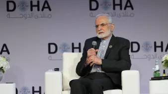 در این تصویر، فردی بزرگسال با ریش و موهای خاکستری در حال نشستن بر روی صندلی است. او لباس رسمی به تن دارد و به نظر می‌رسد در یک نشست یا کنفرانس شرکت کرده است. در پس‌زمینه، لوگوی "دوحه" و متن عربی به چشم می‌خورد که نشان‌دهنده برگزاری یک همایش یا فوروم است. فضای کلی تصویر به صورت مدرن و رسمی طراحی شده و روی میز کنار او، چند بطری آب و گل‌های تزئینی قرار دارد.