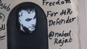 L'image représente un graffiti sur un mur. On y voit un portrait en noir et blanc d'un homme, accompagné d'un texte écrit en anglais. Le message indique "Freedom For HR Defender" (Liberté pour le défenseur des droits de l'homme) et mentionne un nom d'utilisateur Twitter "@nabeelrajab". Le style artistique est simple, avec un fond sombre et des lettres noires qui ressortent sur la surface claire du mur. Cela semble être une œuvre engagée, soulignant le soutien à la défense des droits humains.