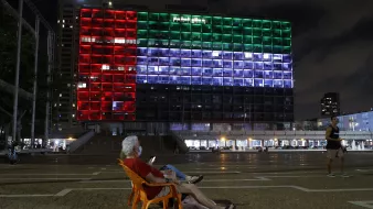 The image shows a large building illuminated with colorful lights at night, displaying the colors of a flag: red, green, and white. In the foreground, an elderly man is sitting in a plastic chair, looking at his phone. The scene conveys a sense of tranquility, with the illuminated building serving as a striking backdrop in a public square. In the background, other people can be seen walking or engaging in activities. The atmosphere appears calm and reflective.