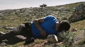 Nell'immagine si vede un uomo sdraiato su un prato verde, con il viso rivolto verso il terreno. Indossa una maglietta blu e pantaloni scuri. Accanto a lui ci sono alcune rocce. Sullo sfondo, si scorge un bulldozer che lavora su un terreno collinare. La scena trasmette un senso di tranquillità, ma anche di inquietudine a causa della presenza del macchinario che sottolinea il contrasto tra l'uomo e l'ambiente circostante.