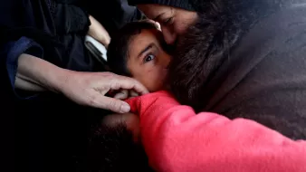L'immagine mostra un momento intimo e toccante tra un adulto e un bambino. Il bambino, con uno sguardo curioso e attento, è avvolto in un abbraccio mentre l'adulto, probabilmente una madre o una figura di riferimento, lo bacia affettuosamente. Le mani di un'altra persona si intravedono, creando una sensazione di protezione e vicinanza. L'atmosfera trasmette emozione e calore umano, evidenziando il legame profondo tra le persone. La scena è caratterizzata da toni scuri e un'illuminazione morbida, che contribuiscono a enfatizzare la tenerezza del momento.
