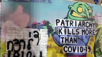 L'image représente une fresque murale colorée et expressive. On y voit un personnage caricatural avec une tête de monstre qui semble joyeux, tenant une pancarte sur laquelle il est écrit "PATRIARCHY KILLS MORE THAN COVID-19". À côté, il y a un graffiti en noir qui semble être écrit dans une autre langue. L'ensemble de l'œuvre rassemble des éléments de critique sociale et politique, mêlant humour et un message sérieux sur le patriarcat et la pandémie.