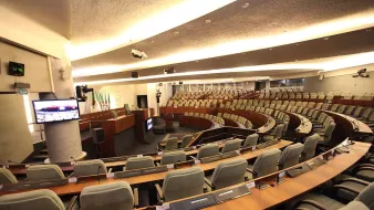 L'image montre une salle de réunion ou un hémicycle, probablement utilisée pour des sessions parlementaires ou des assemblées. On peut voir des rangées de sièges confortables disposés en arc de cercle, avec des tables en bois devant chaque siège. Des écrans et des équipements audiovisuels sont visibles, indiquant que la salle est équipée pour des présentations ou des débats. Les murs sont décorés de manière sobre et moderne, créant une ambiance institutionnelle.