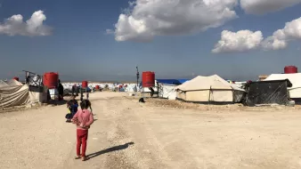 L'image montre un camp de réfugiés avec des tentes disposées de part et d'autre d'un chemin de terre. Au fond, on aperçoit des nuages blancs dans un ciel bleu. Des enfants sont présents, certains semblent jouer ou se déplacer dans le camp. Les abris sont principalement en toile, avec quelques réservoirs d'eau visibles. L'atmosphère paraît calme malgré les conditions de vie apparentes de ces lieux.