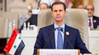 L'image montre un homme assis derrière un bureau lors d'une réunion officielle. Il est habillé en costume sombre et semble sérieux. À côté de lui, il y a un drapeau de la Syrie et une plaque indiquant "République arabe syrienne". En arrière-plan, on peut apercevoir d'autres personnes et une disposition formelle typique d'une conférence ou d'un sommet.