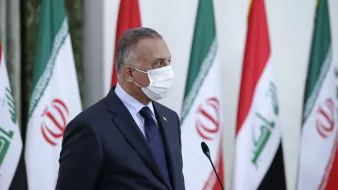 The image depicts a person wearing a mask, standing in profile. He is dressed in a dark suit with a tie and appears to be engaged in an official event. In the background, multiple flags are visible, including those of Iran and Iraq, suggesting a diplomatic or governmental setting. The person appears serious and focused, possibly addressing an audience or participating in a press conference.