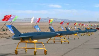 The image depicts a row of military drones lined up on a tarmac. Each drone is mounted on a yellow stand and features colorful flags attached to poles, which are positioned behind them. The background shows a vast, open landscape with distant mountains under a clear sky, suggesting a desert environment. The overall scene conveys a sense of military readiness or demonstration.