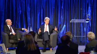 L'image montre une scène de conférence ou de discussion, où deux intervenants sont présents sur une scène. À gauche, un homme est assis sur un fauteuil, écoutant attentivement, tandis qu'à droite, un autre homme parle avec des gestes, probablement en train de partager ses idées ou répondre à des questions. La scène est éclairée avec un fond de rideaux bleus, créant une ambiance professionnelle et formelle. Il y a également un podium avec un microphone, prêt à être utilisé. En bas, on peut apercevoir un public assis, attentif à la conversation.