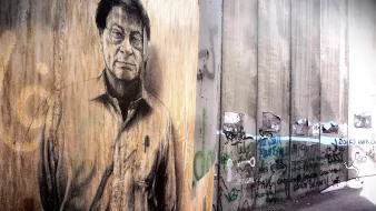 L'immagine rappresenta un murale su un muro di cemento, caratterizzato da un ritratto di un uomo con occhiali e un'espressione seria. Il muro è grezzo e presenta anche scritte e graffiti, suggerendo un contesto urbano e di protesta. Il contrasto tra il ritratto e il resto del muro evidenzia un messaggio artistico e sociale. La scena evoca un senso di riflessione e di critica.