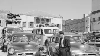L'image montre une scène de rue animée, probablement dans un contexte urbain d'une époque passée. Au premier plan, un homme en costume se tient debout, l'air décontracté, avec un pied posé sur une petite caisse. Devant lui, un autre homme est accroupi, occupé à préparer quelque chose dans un petit pot ou une casserole. En arrière-plan, on peut voir de nombreuses voitures anciennes, avec des bagages sur leur toit, suggérant peut-être un moment de transition ou de voyage. Le cadre est lumineux et semble capter une ambiance d'activité dynamique, typique des villes dans les décennies passées.