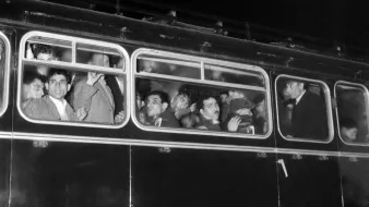 L'image montre un bus noir en soirée, dont les fenêtres sont remplies de passagers. On peut voir plusieurs visages, certains semblent surpris ou curieux, tandis que d'autres affichent des expressions neutres. L'éclairage est faible, accentuant l'atmosphère nocturne de la scène. Le bus a l'air ancien, avec une carrosserie en métal et des détails rétro.