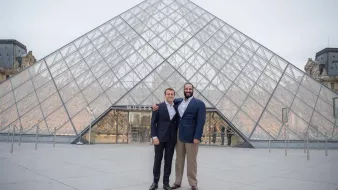 L'image montre deux hommes posant ensemble devant le Musée du Louvre, qui est célèbre pour sa pyramide en verre. Le bâtiment est moderne et impressionnant, contrastant avec l'architecture classique des ailes du musée. Les hommes sourient et portent des costumes formels, se tenant côte à côte, tandis que le ciel est légèrement nuageux, ajoutant une atmosphère calme à la scène.