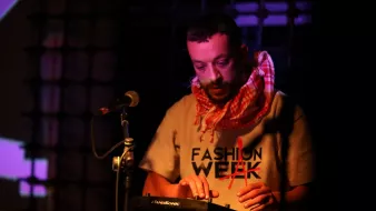 L'immagine mostra un musicista che sta esibendosi su un palco. Indossa una maglietta con la scritta "FASHION WEEK" e una sciarpa a quadri. È concentrato mentre suona un dispositivo elettronico, probabilmente un sintetizzatore o una drum machine. Il palco è illuminato da luci colorate che creano un'atmosfera vivace e coinvolgente. Dietro di lui, si intravedono proiezioni visive che aggiungono dinamismo alla performance.