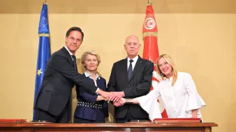 En la imagen se puede ver a cuatro personas realizando un apretón de manos en un ambiente ceremonial. Dos banderas, una de la Unión Europea y otra de Túnez, están visibles en el fondo. Los participantes parecen ser líderes políticos, y todos lucen serios y atentos. La habitación tiene un diseño formal, lo que sugiere que se trata de un evento importante o una firma de acuerdo.