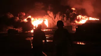 The image depicts two silhouetted figures walking towards a large fire at night, with bright flames and thick smoke rising in the background. The scene conveys a sense of danger and urgency.